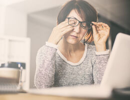 A woman rubs her eye while working at a laptop